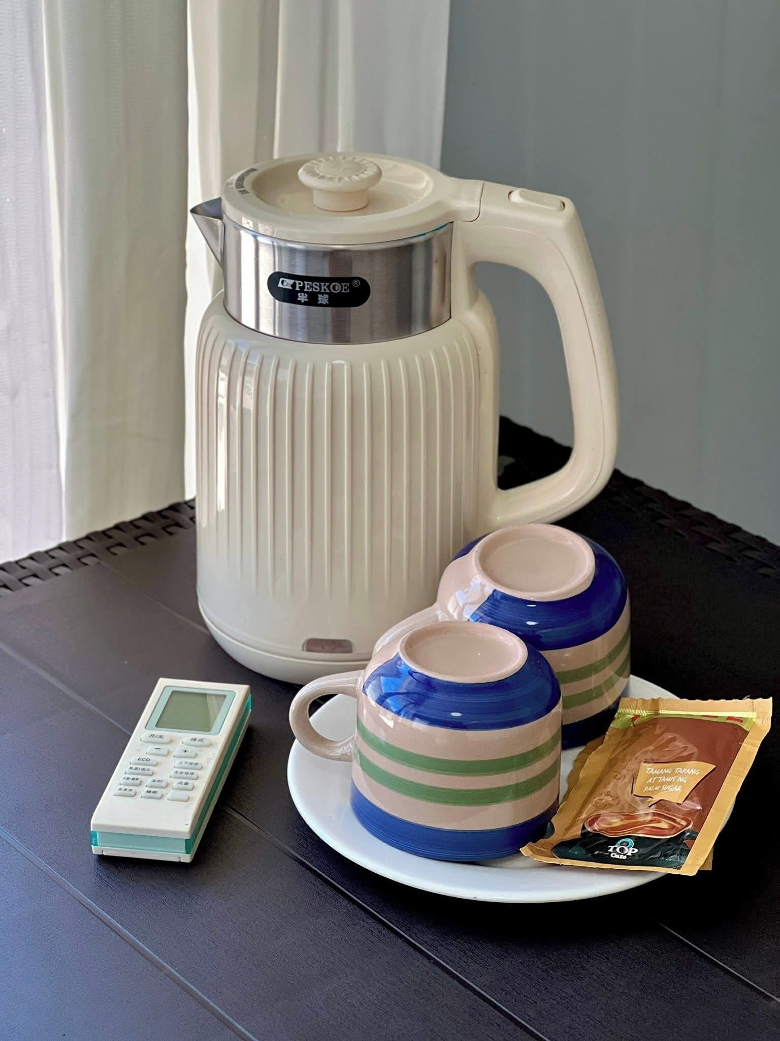A white electric kettle, two striped cups, a small digital device, and a packet of tea on a table.