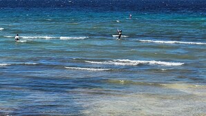 Una playa con cielos despejados, dos cometas volando y una persona practicando surf.