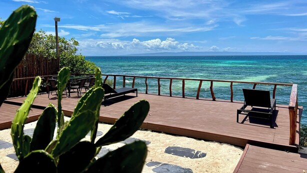 Terraza de madera con sillas que da a un mar turquesa, una cerca de madera y una planta de cactus en primer plano.