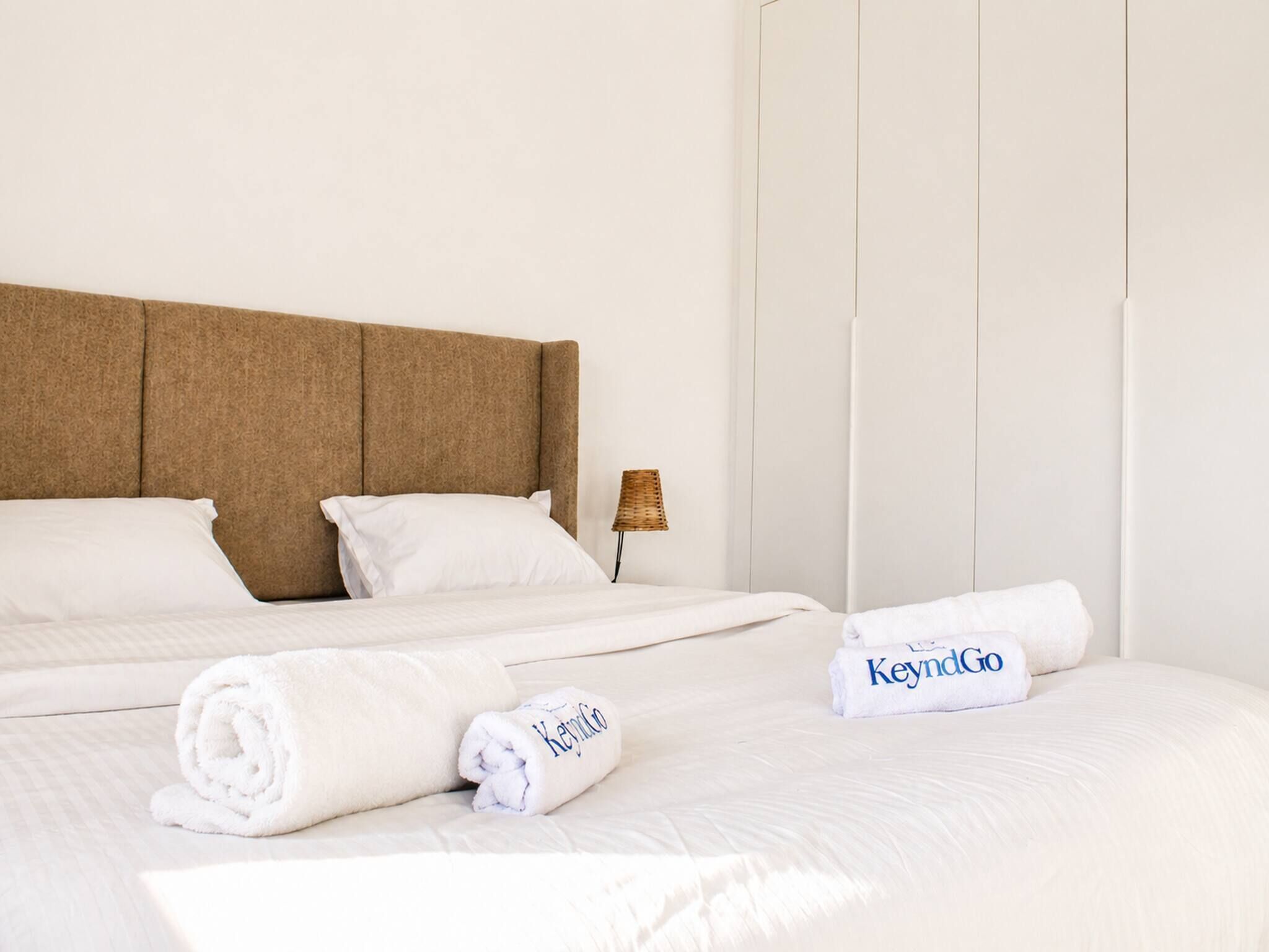 Een net opgemaakt bed met witte lakens, een bruin hoofdbord en twee handdoeken met 'KeyndGo'-branding.