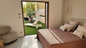 Una camera da letto con un letto, una sedia, un tavolino e vista su un giardino con un albero e un lettino.