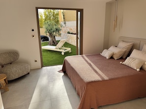 Ein Schlafzimmer mit Bett, einem Sessel, einem kleinen Tisch und Blick auf einen Garten mit Baum und Liegestuhl.