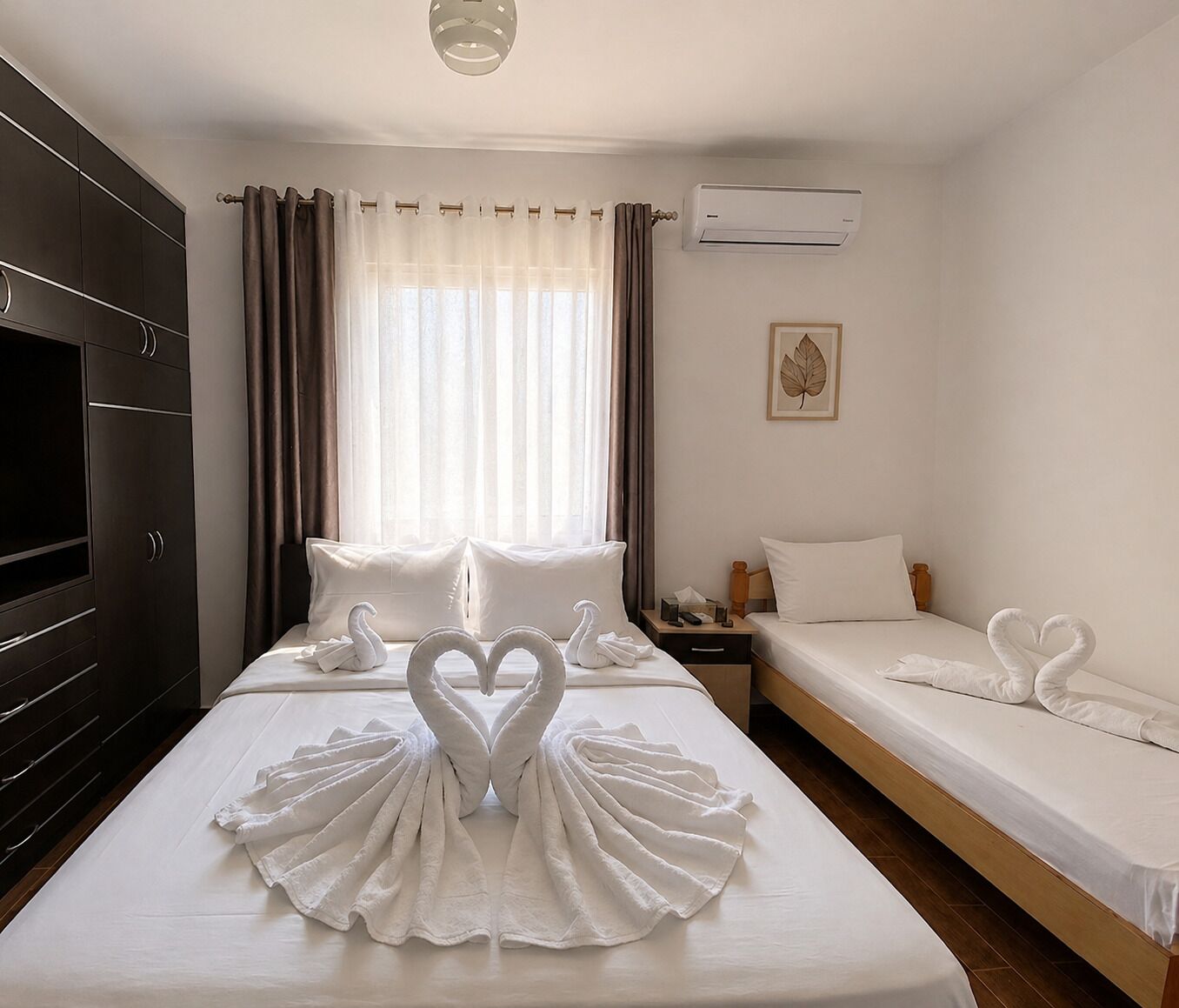 A hotel room with a large bed, a smaller bed, a wardrobe, an air conditioning unit, and a window with curtains.