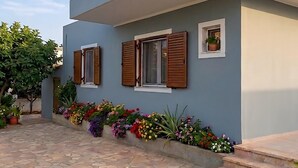 Un bâtiment moderne avec une entrée pavée de pierres, des volets en bois aux fenêtres et une variété de fleurs colorées le long du chemin.