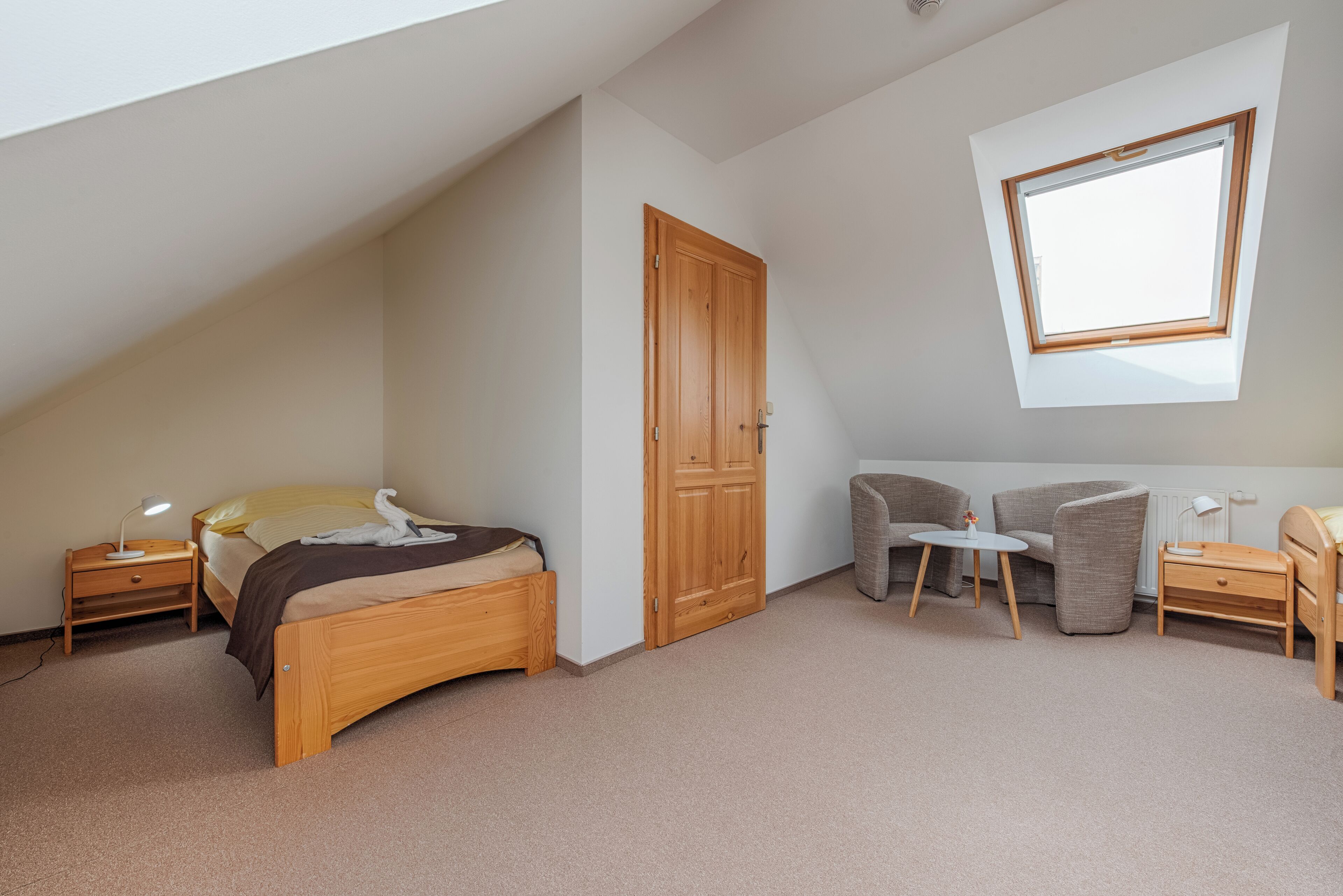 A bedroom with a sloped ceiling, a wooden wardrobe, a bed with a blanket, a small table with a lamp, and a window.
