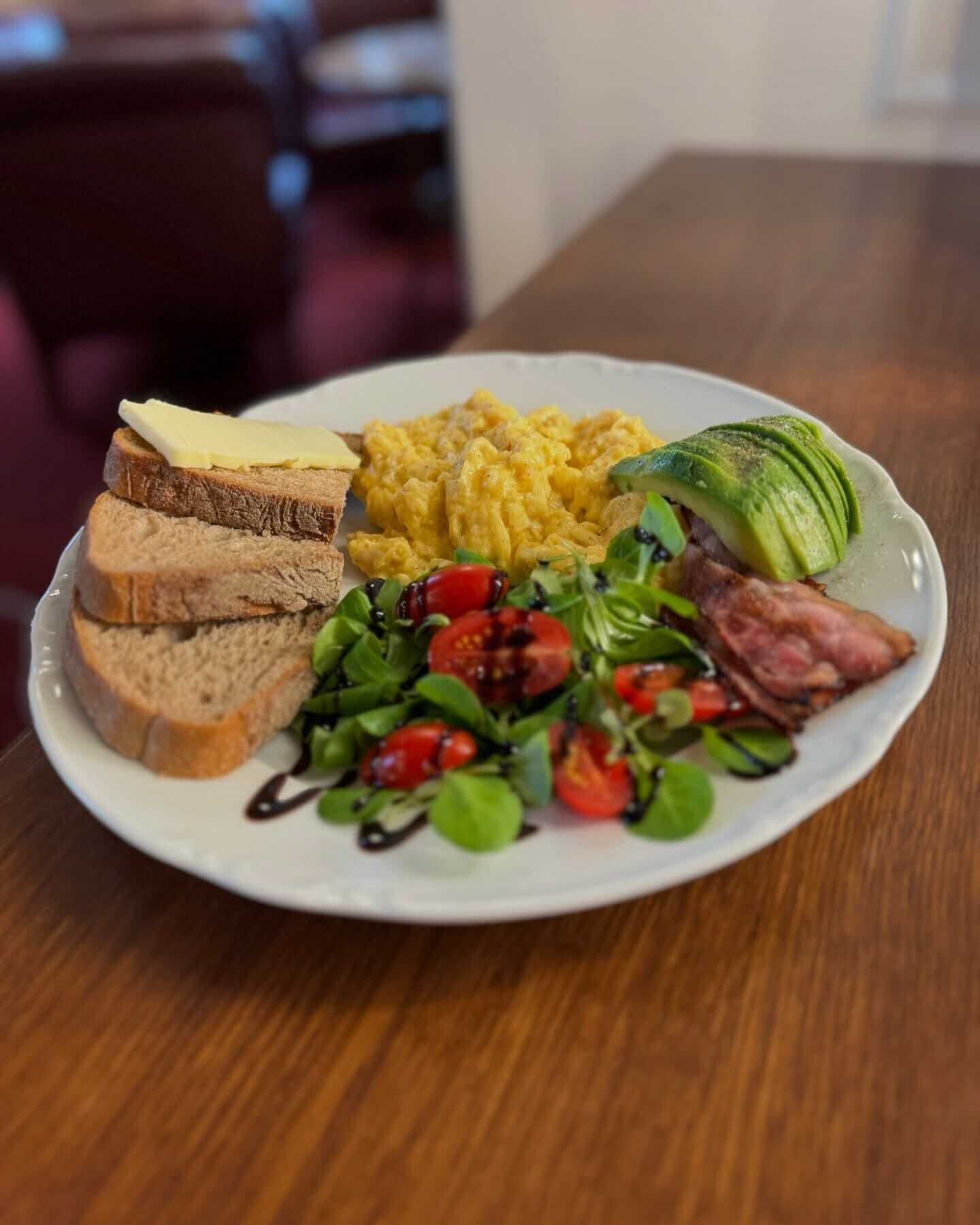 A plate with scrambled eggs, avocado slices, a meat dish, a salad, and toast with butter on a wooden table.