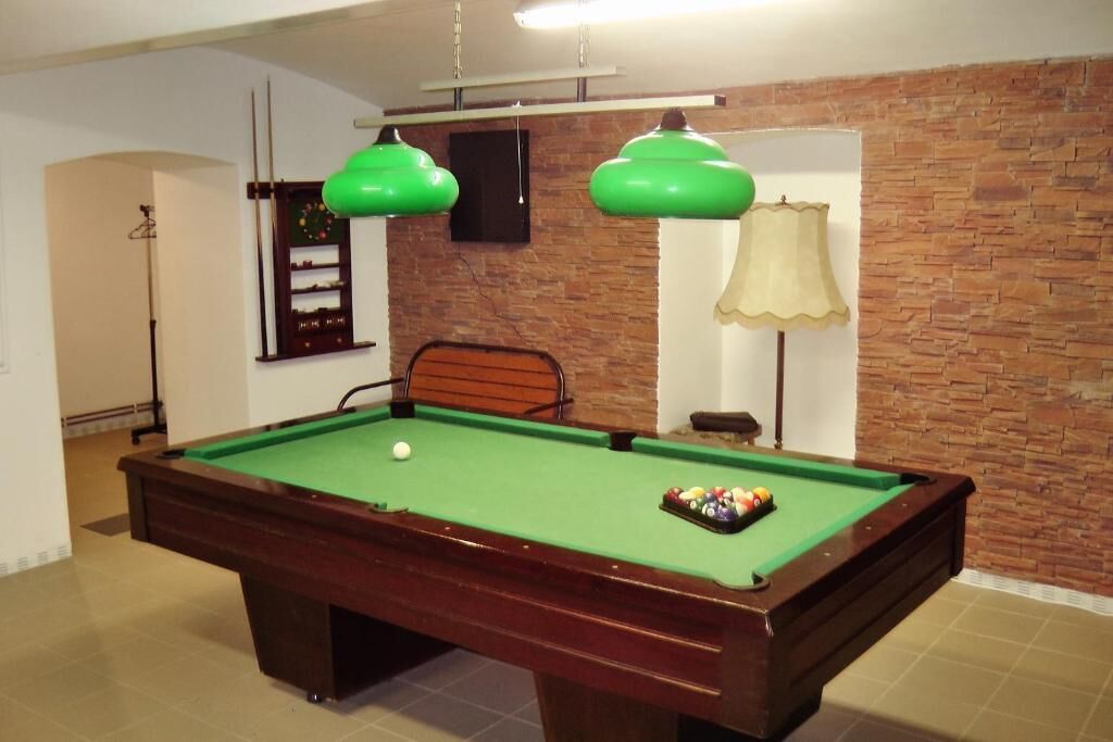 Una mesa de billar con superficie de fieltro verde, una bola blanca y un juego de bolas de billar. Dos lámparas colgantes verdes están encima de la mesa, una lámpara con pantalla beige y una pared con diseño de ladrillo.