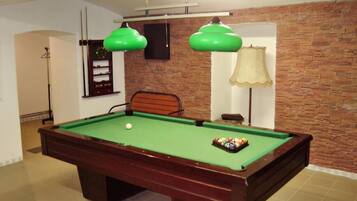 Una mesa de billar con superficie de fieltro verde, una bola blanca y un juego de bolas de billar. Dos lámparas colgantes verdes están encima de la mesa, una lámpara con pantalla beige y una pared con diseño de ladrillo.