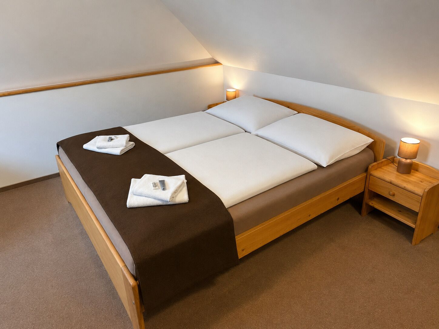 A single bed with a brown headboard, white bedding, and two towels on the bed. There is a wooden nightstand with a lamp beside the bed.