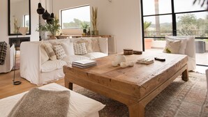 A spacious living room with a large wooden coffee table, white sofas, and a large window offering a view of the outdoors.