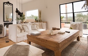 Ein geräumiges Wohnzimmer mit einem großen Holztisch, weißen Sofas und einem großen Fenster mit Blick ins Freie.