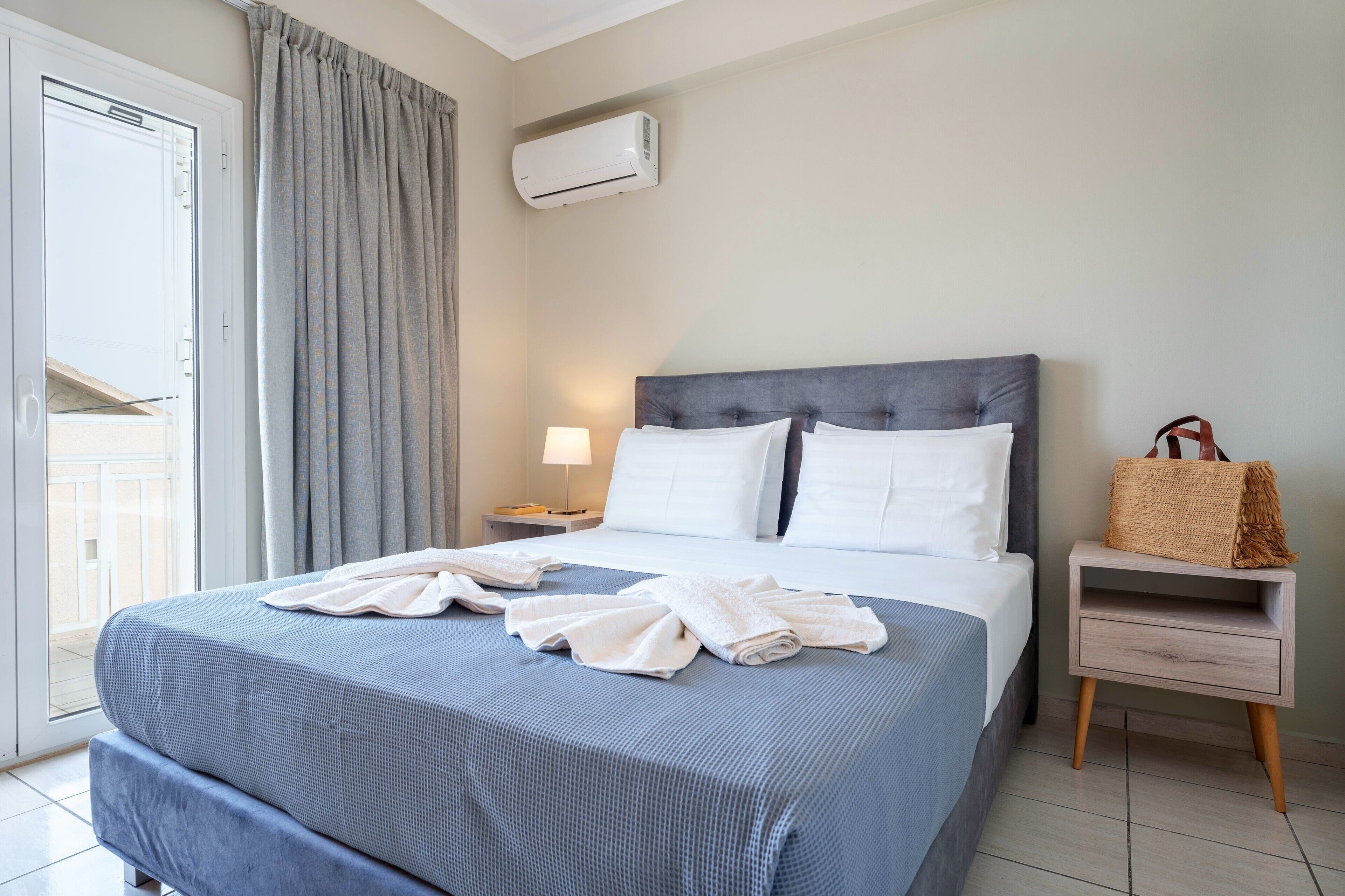 Quarto de hotel com cama, mesa de cabeceira, abajur, mala e aparelho de ar condicionado.
