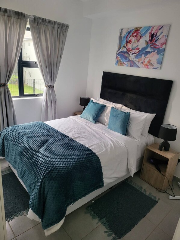 A bedroom with a bed, curtains, a nightstand, a lamp, a painting, and a view of the outside. - Modern Coastal Escape in Salt Rock Ballito - Perfect Family Retreat (Ballito)