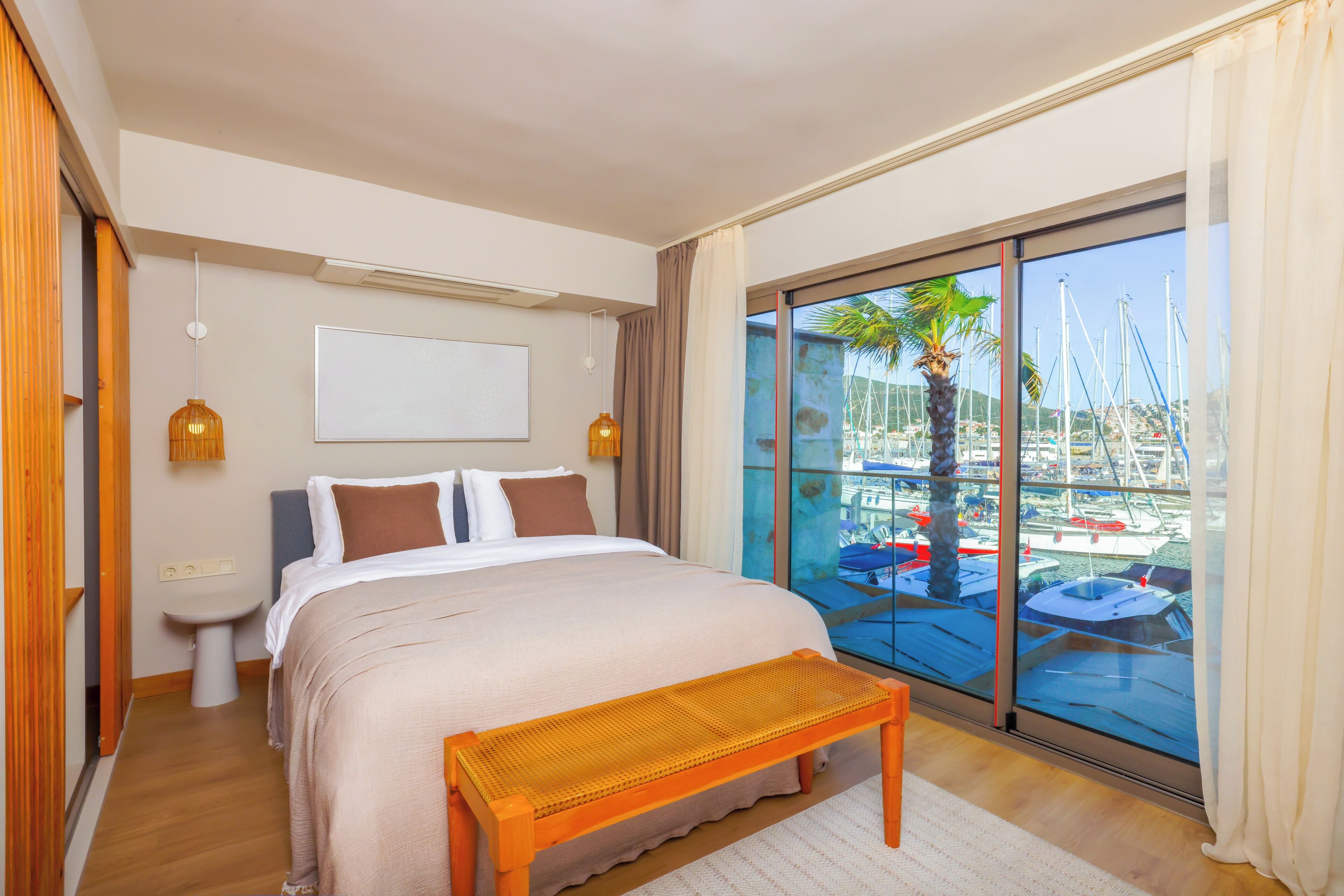 A hotel room with a bed, bedside table, lamp, and a view of boats through a large window.