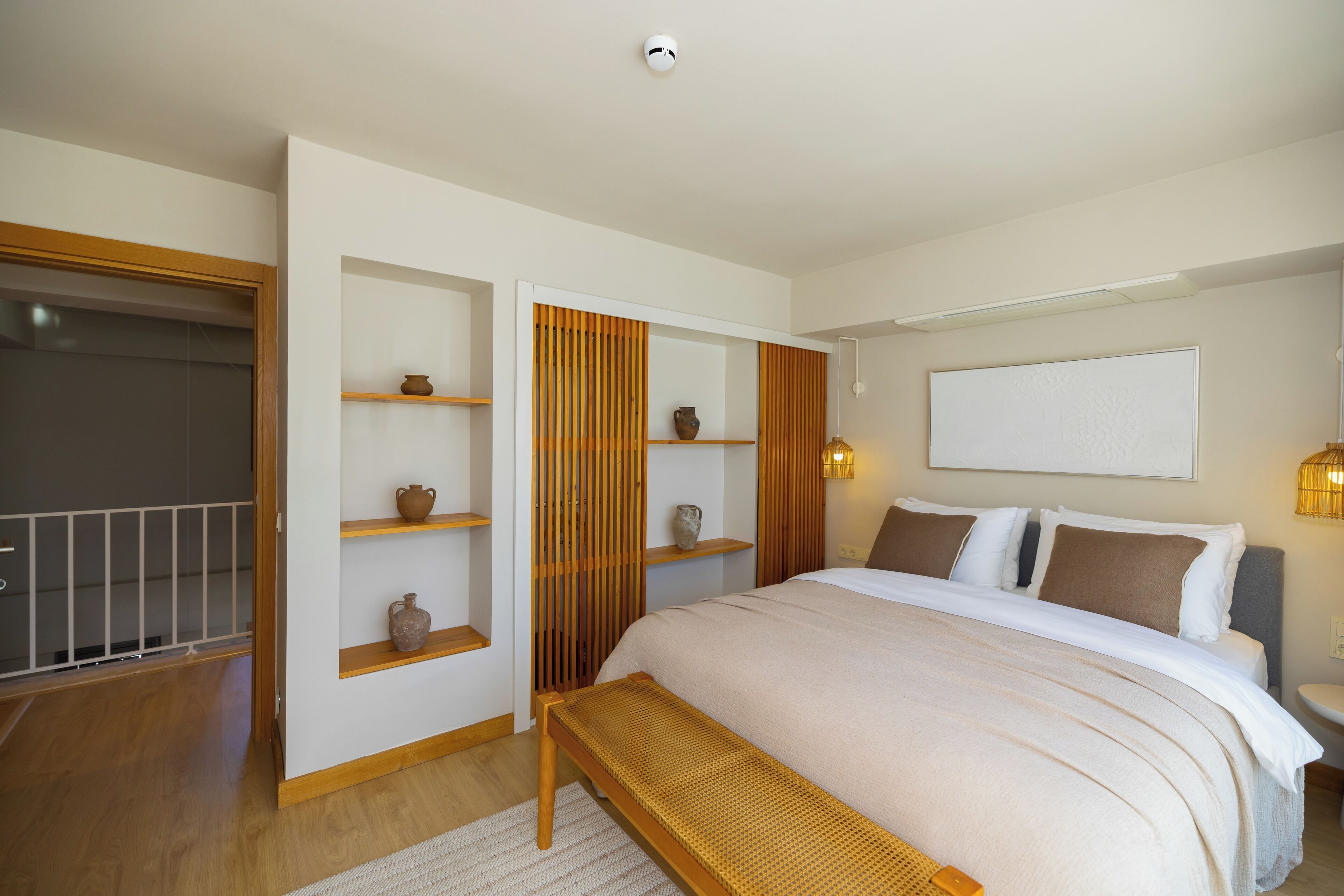 A bedroom with a bed, wooden shelves, a bench, a wardrobe, and a bedside table with a lamp.