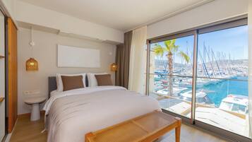 Quarto com cama grande, mesa de cabeceira, um banco, vista para uma marina com barcos e paisagem urbana ao longe.