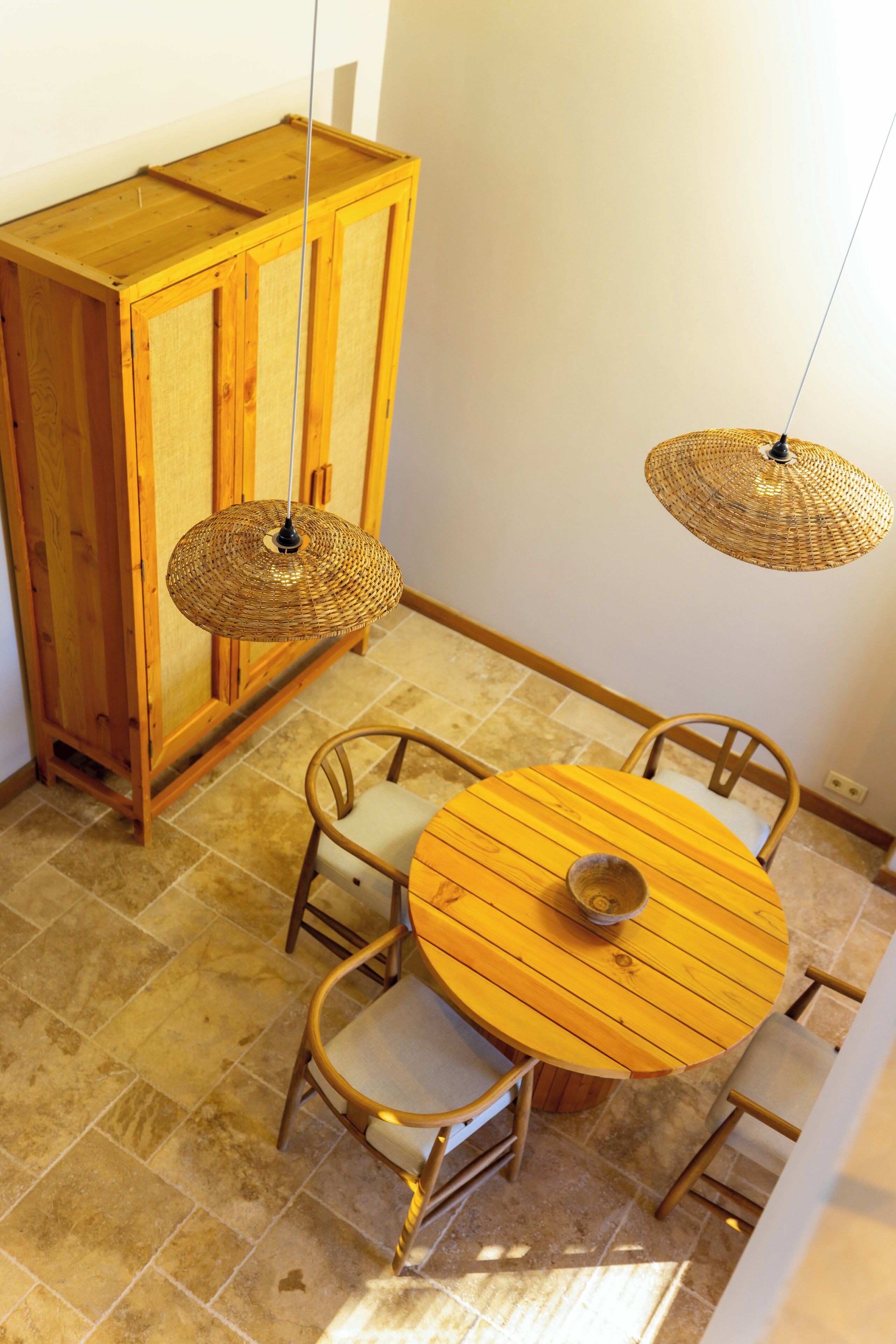 Un coin repas comprenant une table ronde en bois, quatre chaises, un luminaire suspendu en fibres tressées et un meuble en bois.