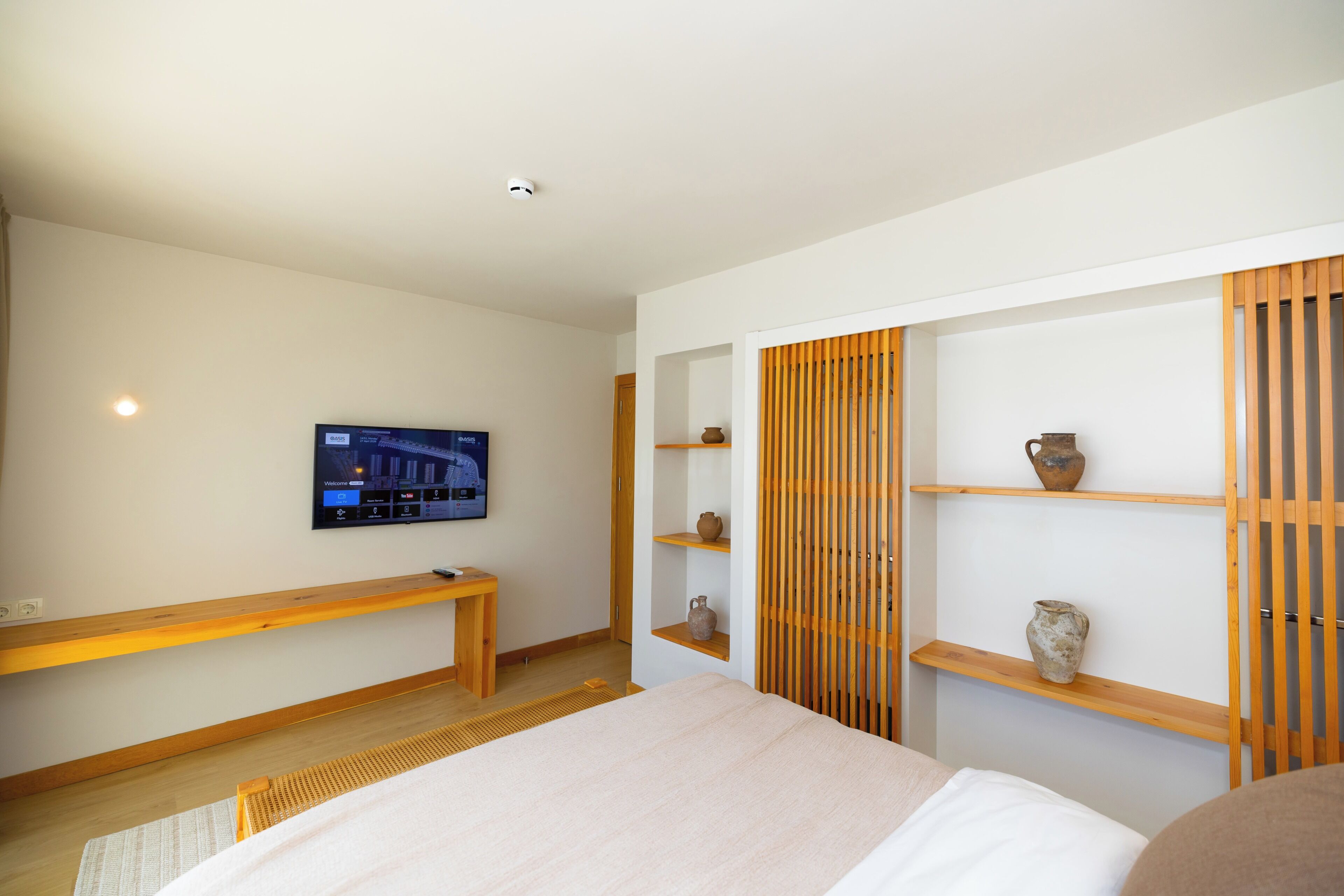 A modern hotel room with a bed, a TV mounted on the wall, wooden shelves with decorative items, and a bench.