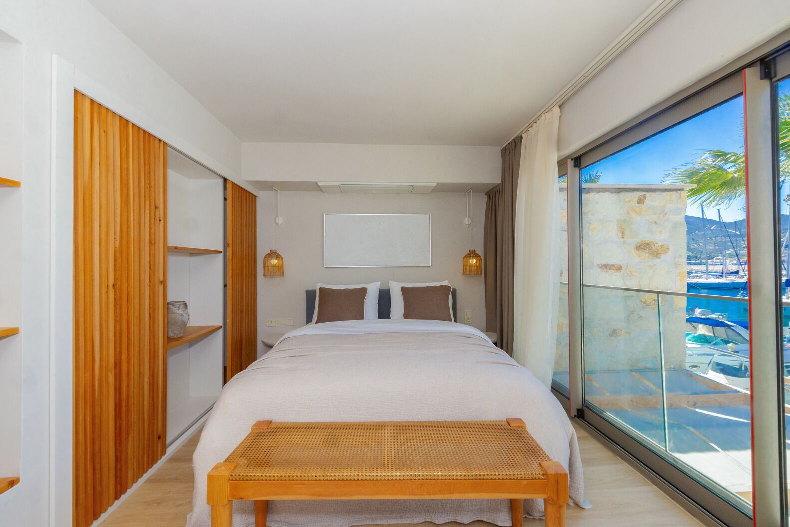 Une chambre moderne avec un grand lit, une armoire en bois et une vue sur une marina, visible à travers une porte vitrée.