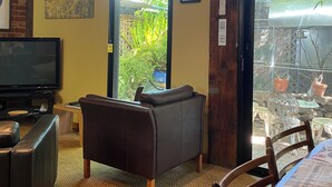 Un salon avec un téléviseur, un canapé en cuir et une table à manger avec des chaises. Des tableaux encadrés ornent le mur, et on peut voir le jardin à travers la porte.