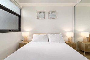 Une chambre d’hôtel avec un lit, deux tables de chevet avec des lampes, une petite plante en pot et deux tableaux encadrés au mur.