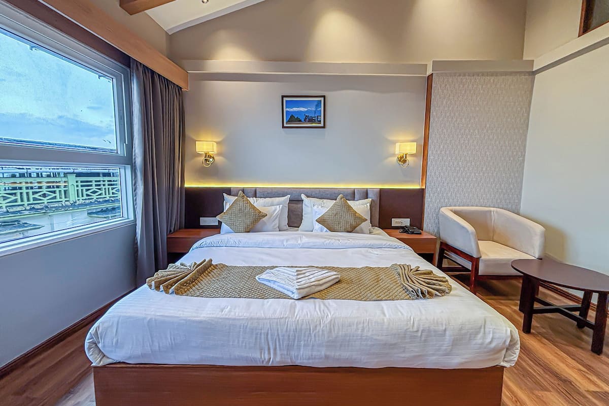 A hotel room with a large bed, a chair, a small table, a nightstand, a lamp, and a window with a view.