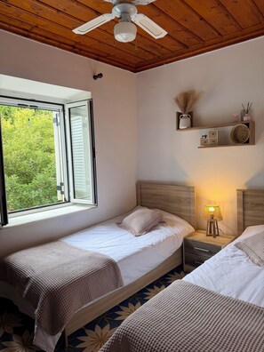 Ein Schlafzimmer mit zwei Betten, einem Deckenventilator, einem Fenster mit Blick ins Grüne, einer Nachttischlampe und einem Regal mit Dekoration.
