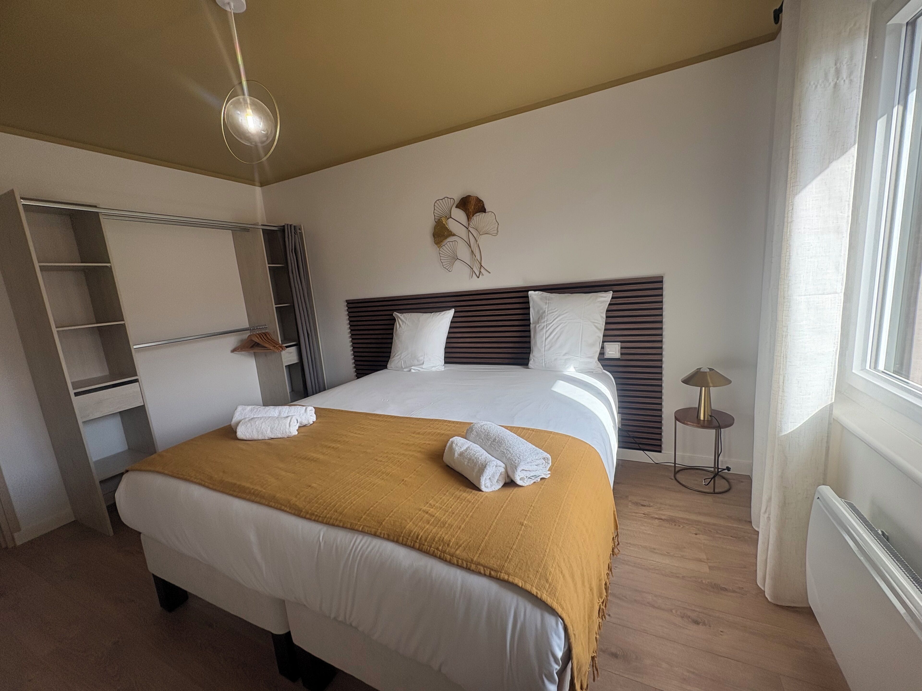 Une chambre d’hôtel comprenant un lit, une lampe de chevet, une fenêtre avec des rideaux, une étagère fixée au mur et un luminaire au plafond.