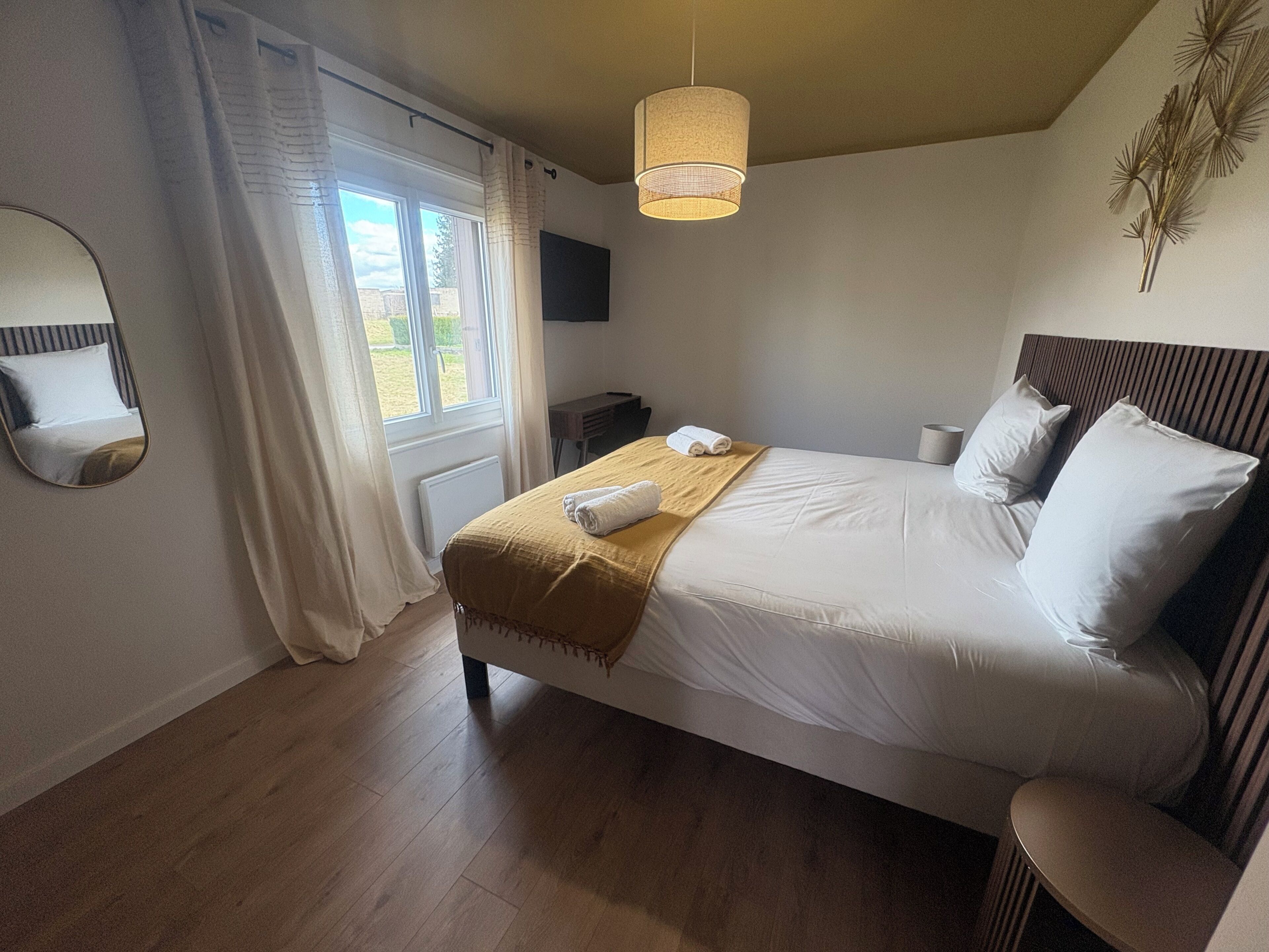 A bedroom with a bed, a bedside table, a mirror, a hanging light fixture, and a window with curtains.