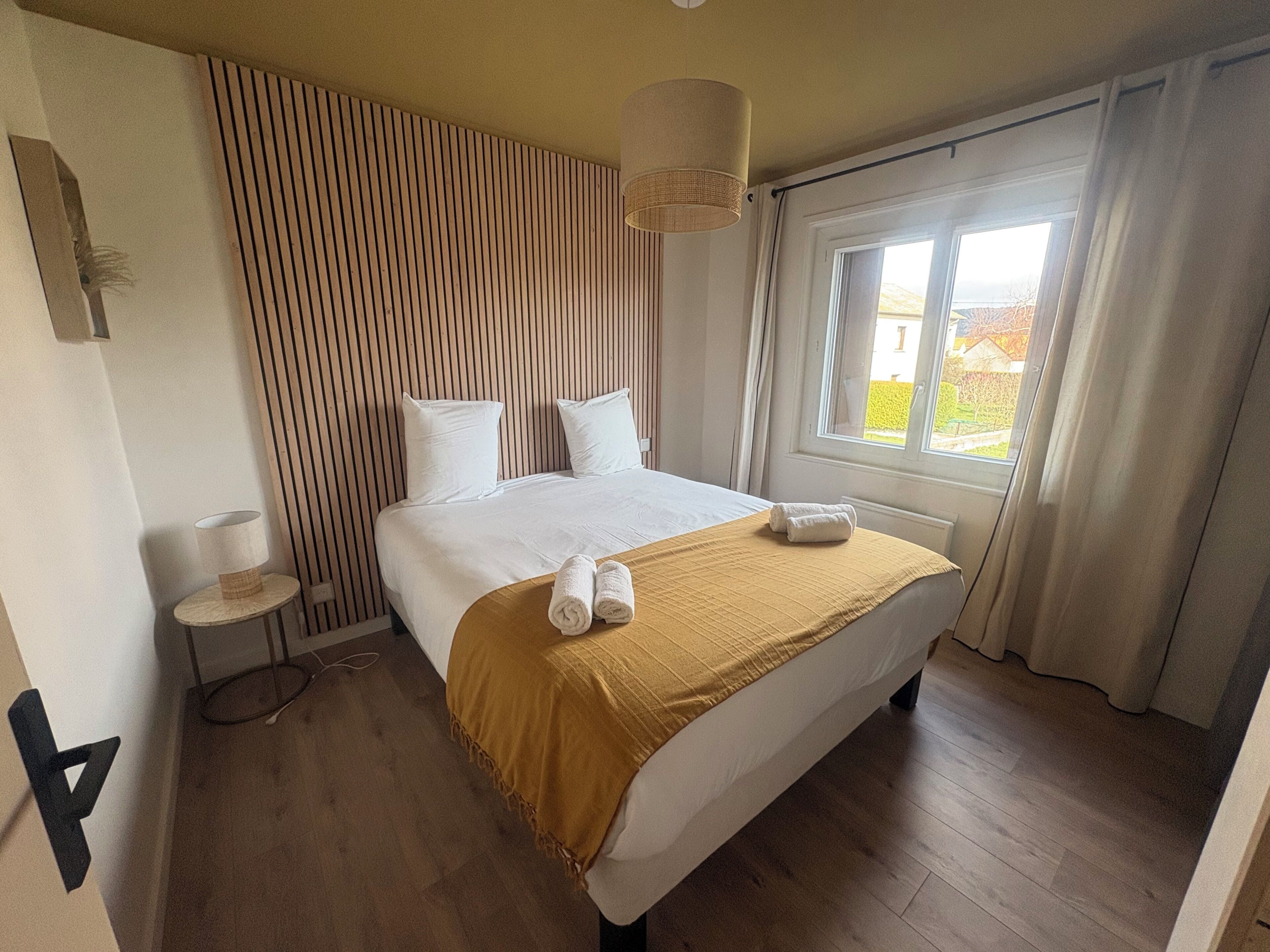 Habitación de hotel con cama, mesita de noche, lámpara, ventana con cortinas y vistas al exterior.