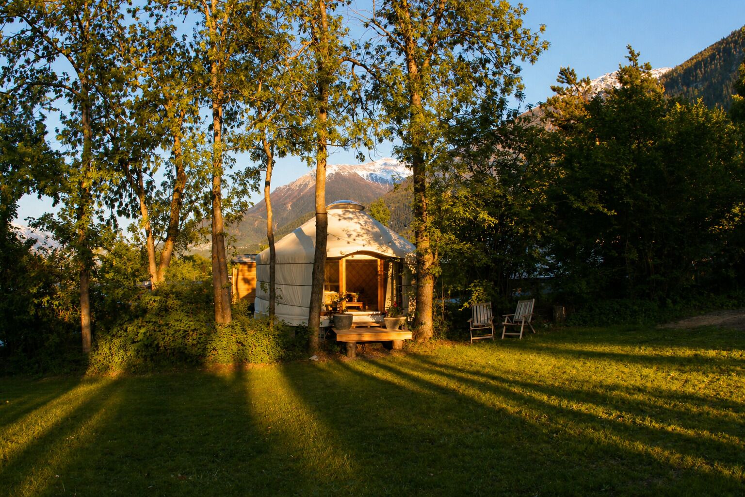 A yurt with a wooden deck and a table, surrounded by trees and a mountain in the background.