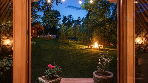 A wooden door opening to a well-lit garden with a fire pit, potted plants, and a wooden deck. - WOODMOOD yurt with wellness (Leuk)