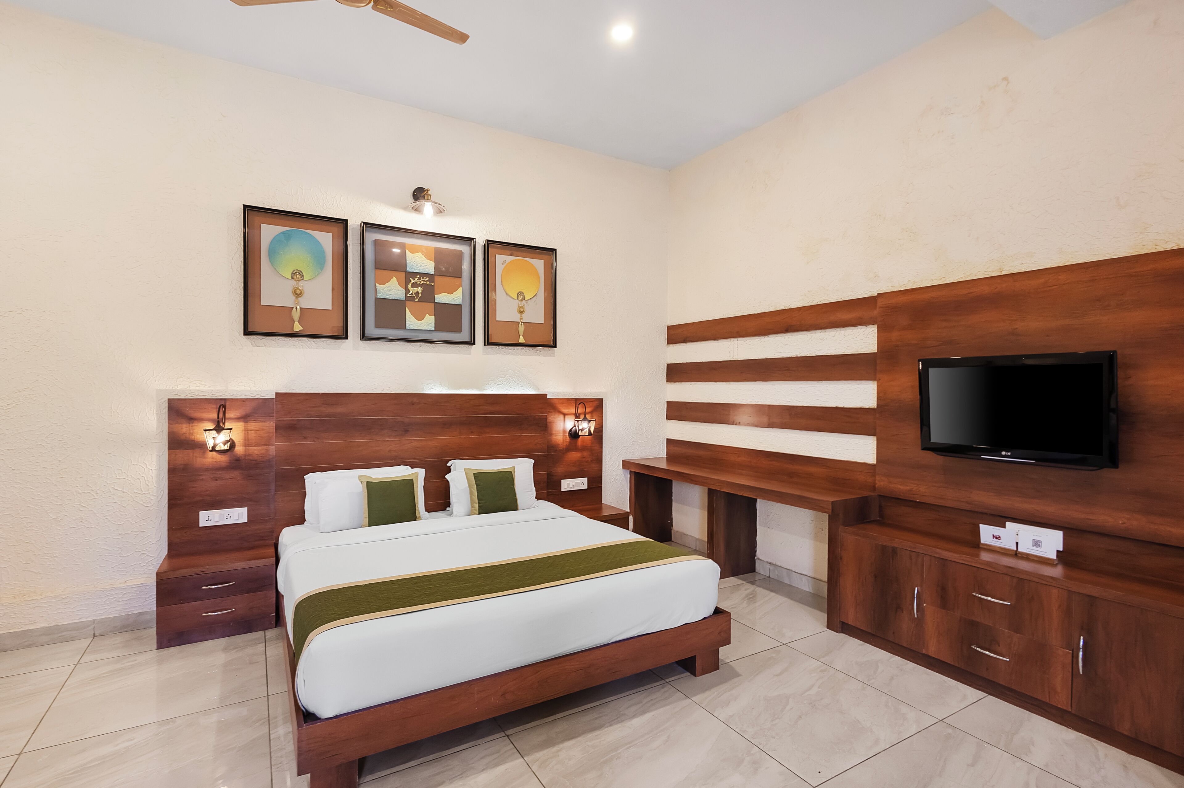 A hotel room with a large bed, wooden headboard, wall-mounted TV, and artwork.