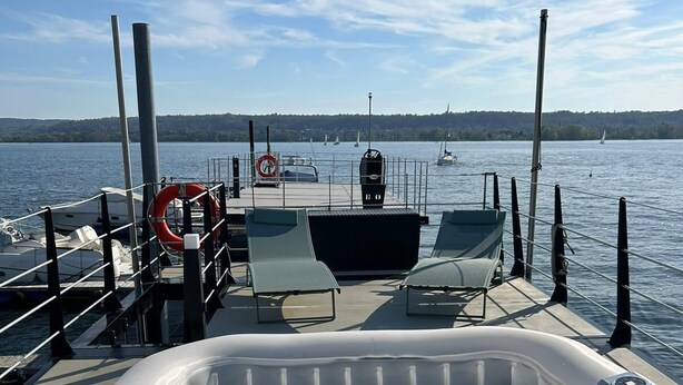 A hot tub on a boat deck with lounge chairs, a lifebuoy, and a view of the water with boats and a distant shoreline. - Celesia Sky water (Sesto Calende)