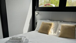 A neatly made bed with white linens and two folded towels, a window with blinds, and a view of greenery outside. - Celesia Sky water (Sesto Calende)