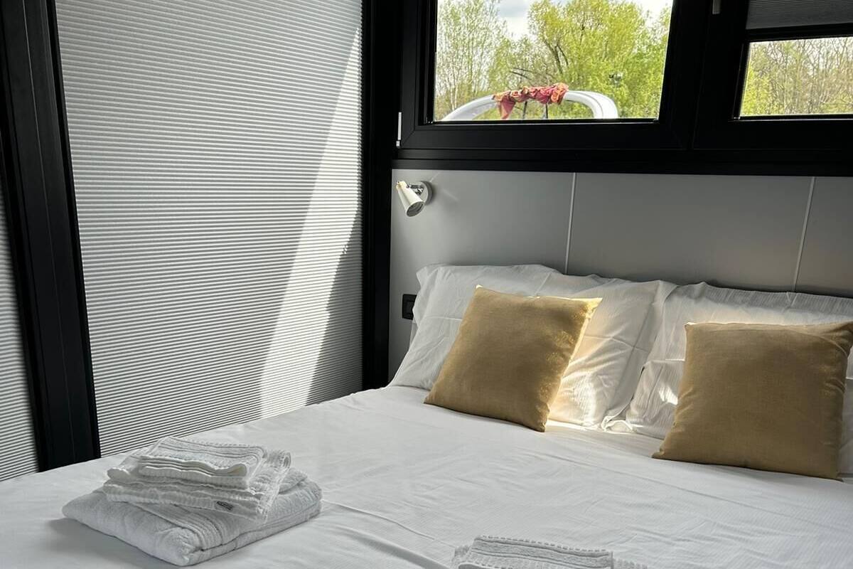 Uma cama bem arrumada com lençóis brancos e duas toalhas dobradas, uma janela com persianas e vista para o verde lá fora.