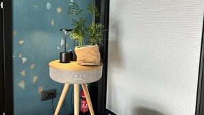 A room with a teal wall, a round wooden table with a plant, a basket with a gray blanket, and a window with blinds. - Celesia Sky water (Sesto Calende)