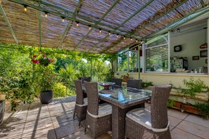 Een terras buiten met rieten meubels, een glazen tafel en potplanten.