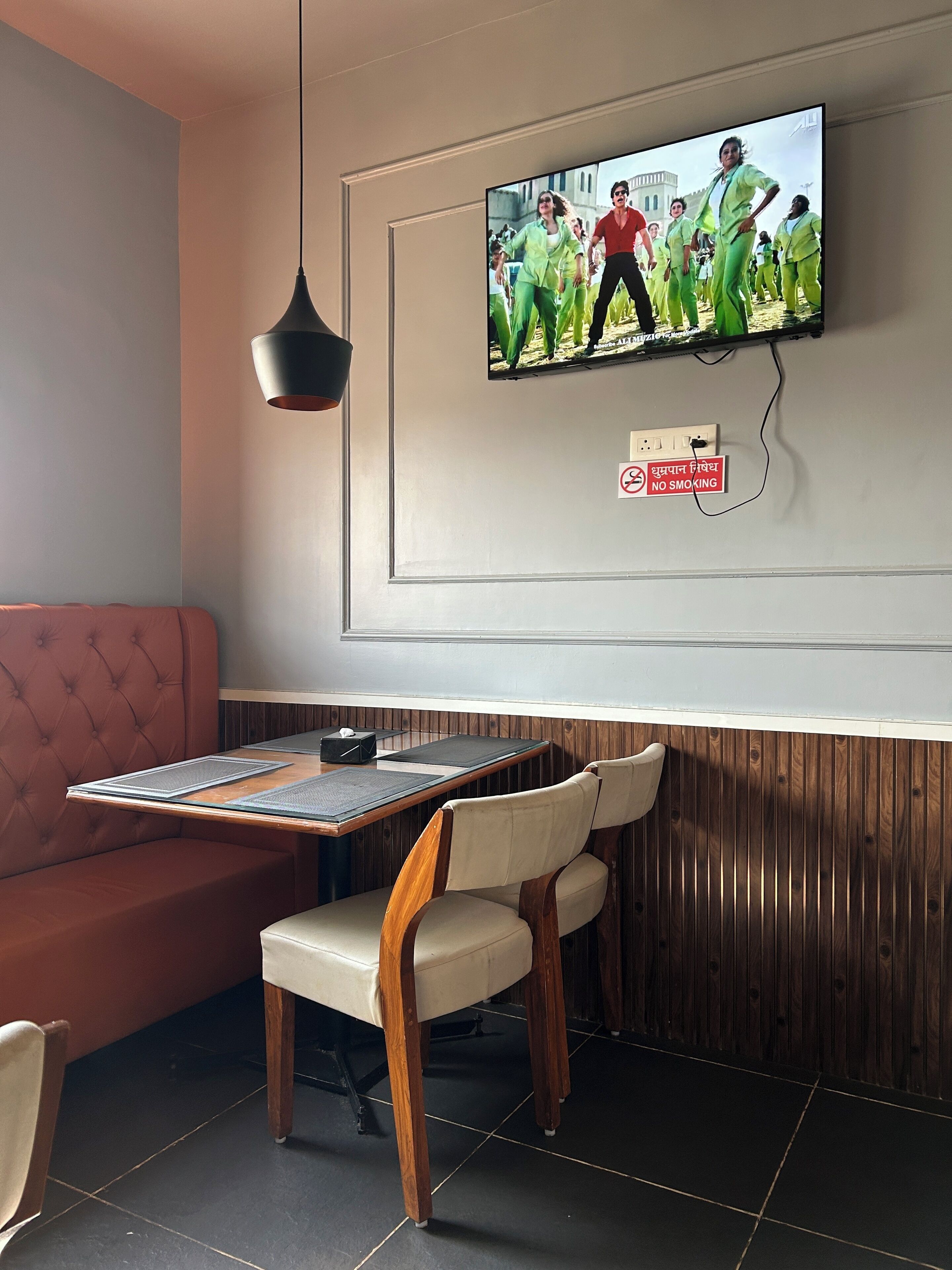 A restaurant interior with a television mounted on the wall displaying a group of people dancing, a 'No Smoking' sign, a cushioned booth, and a wooden table with chairs.