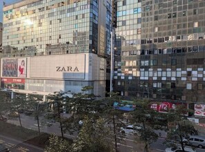 Um edifício moderno com uma loja Zara no andar térreo, grandes janelas de vidro e vista para a rua com árvores e carros estacionados.