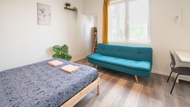 Un dormitorio con una cama, un sofá azul, un escritorio y una ventana con cortinas.