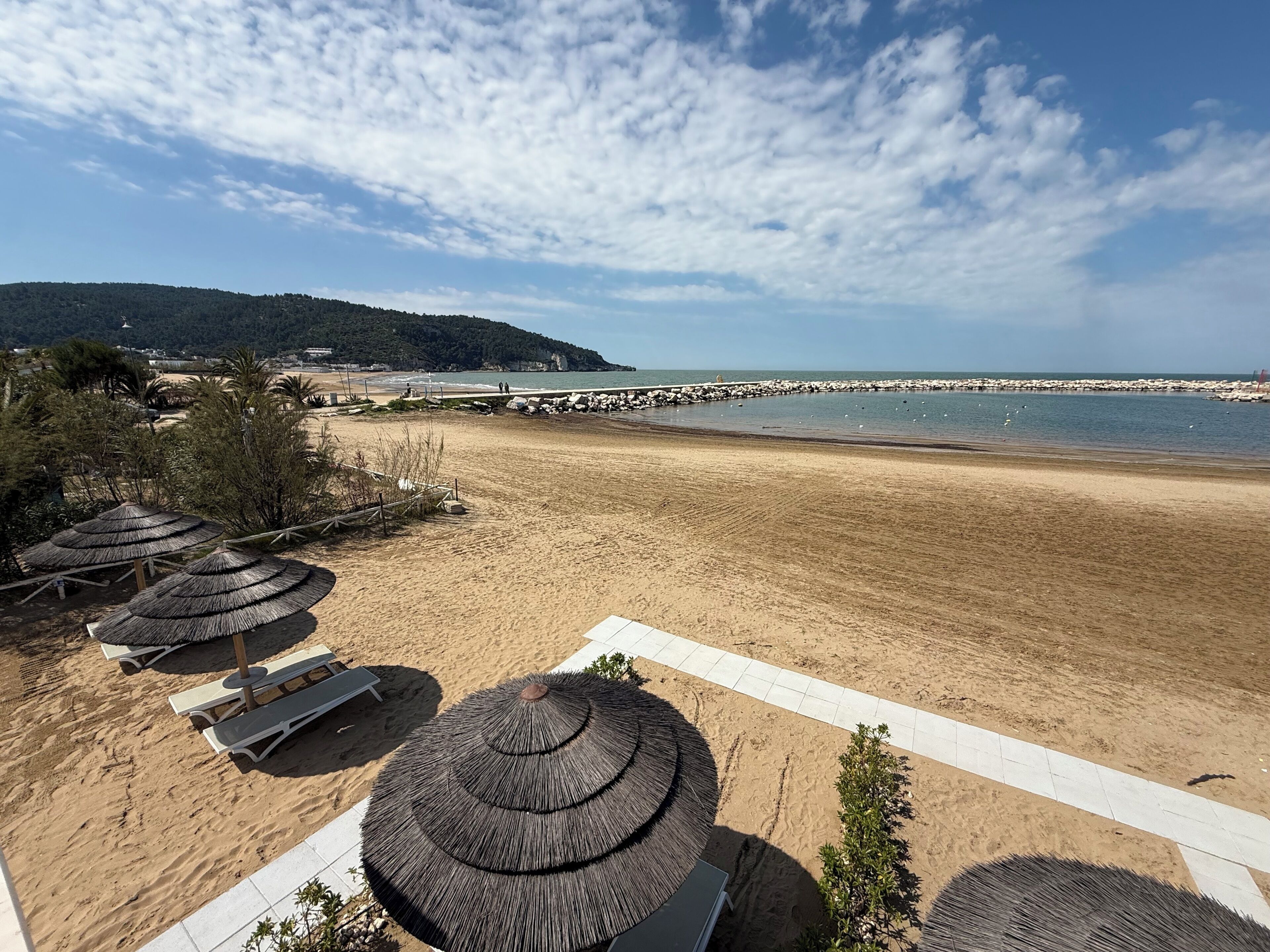 Een strand met parasols en ligbedden, een rotsenrijke golfbreker en uitzicht op de zee en verre heuvels.