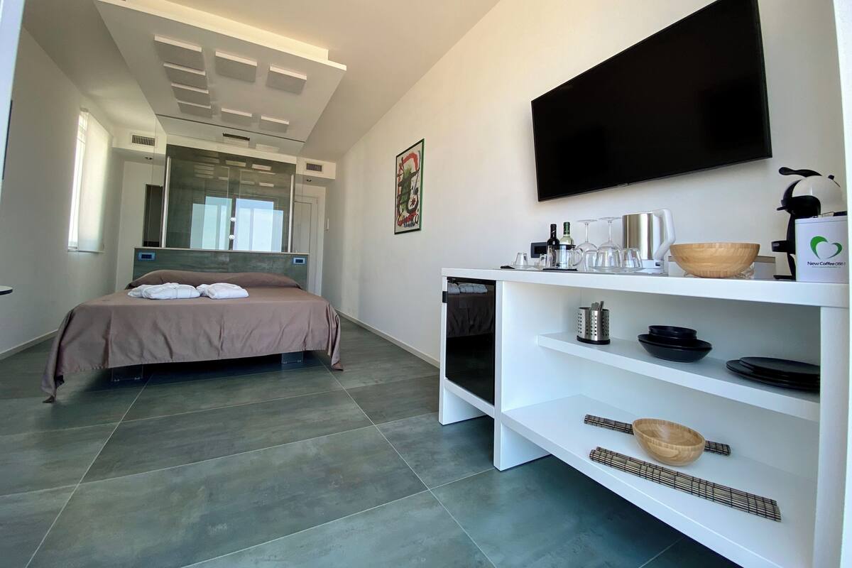 Una habitación de hotel moderna con una cama, un televisor, un estante blanco con utensilios de cocina y un espejo grande.