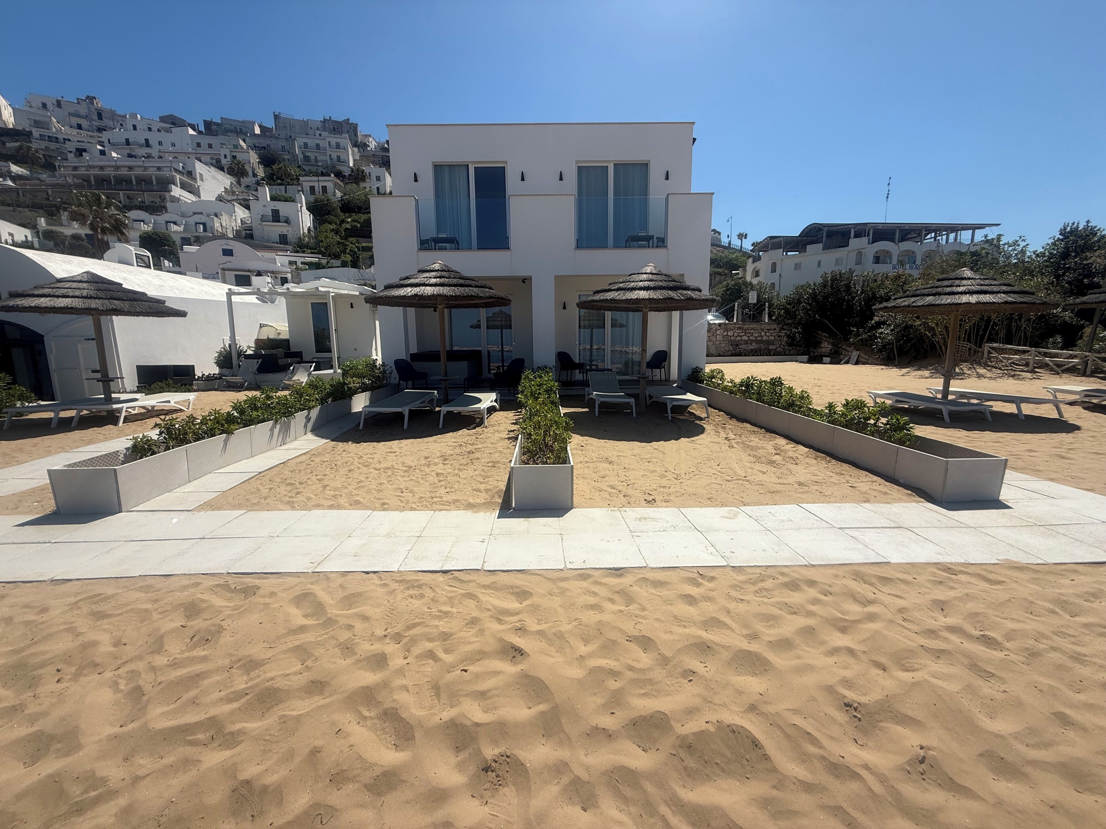 Un hotel moderno con zona de patio que incluye sillas de playa y sombrillas, una playa de arena y vistas de los edificios en las colinas circundantes.