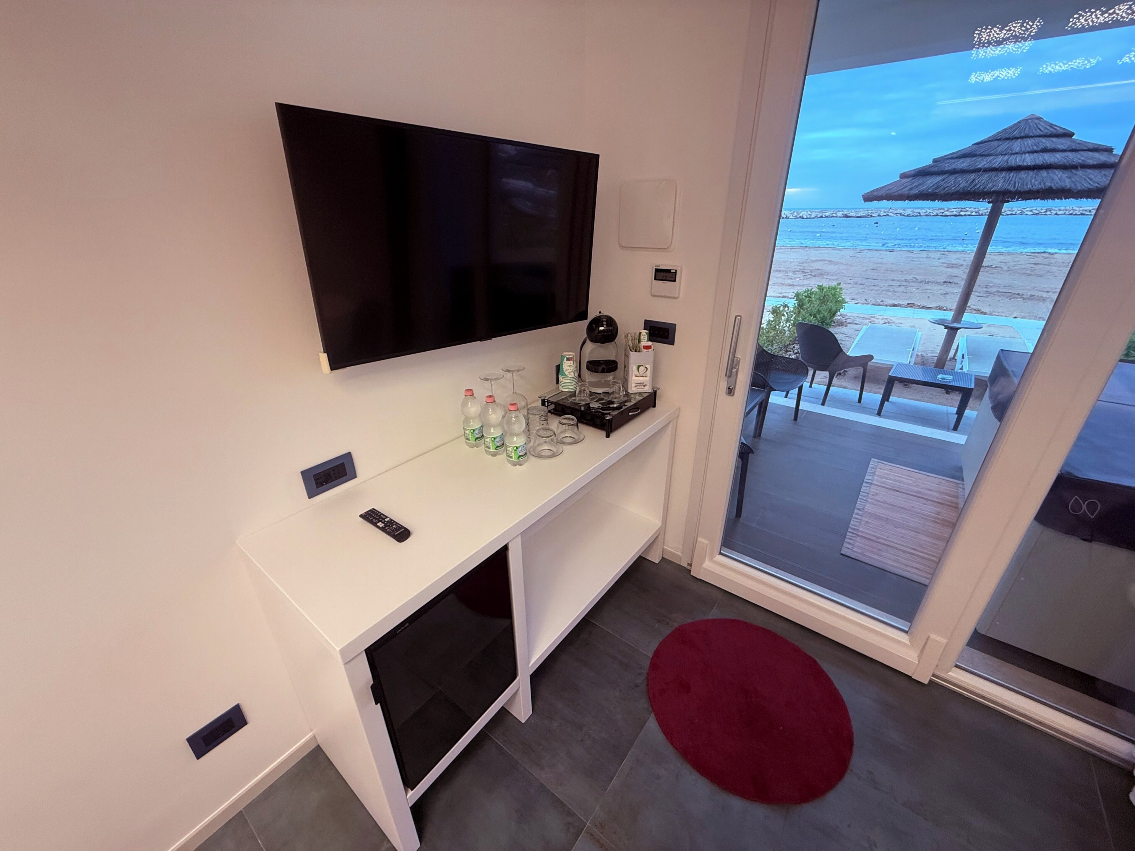 A modern room with a flat-screen TV, a white console table with bottles and glasses, a red rug, and a sliding glass door leading to a balcony with outdoor seating.