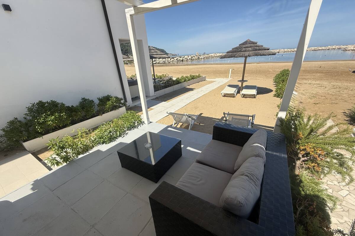 Un espacio exterior moderno con sofás y una mesa, con vistas a una playa con sombrillas y sillas reclinables.