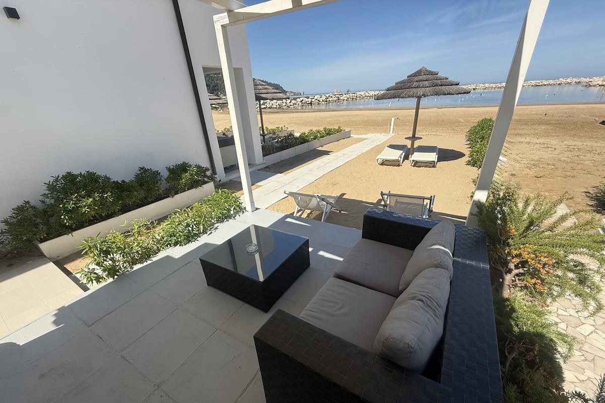 A modern outdoor seating area with a sofa and a table, overlooking a beach with umbrellas and lounge chairs.