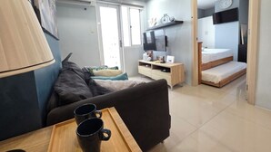 A modern living room with a sofa, a coffee table with two mugs, a TV, a bedside table, and a bed in the background. - Cozy 2-bedroom condo w/AC living room, balcony, Netflix walking dis to SM North (Quezon City)
