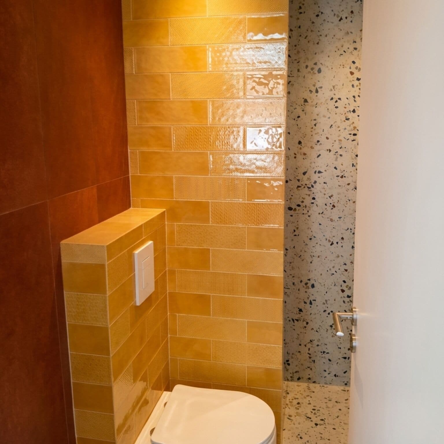 A small, enclosed bathroom with a toilet, a wall-mounted light, and a door with a metallic handle.