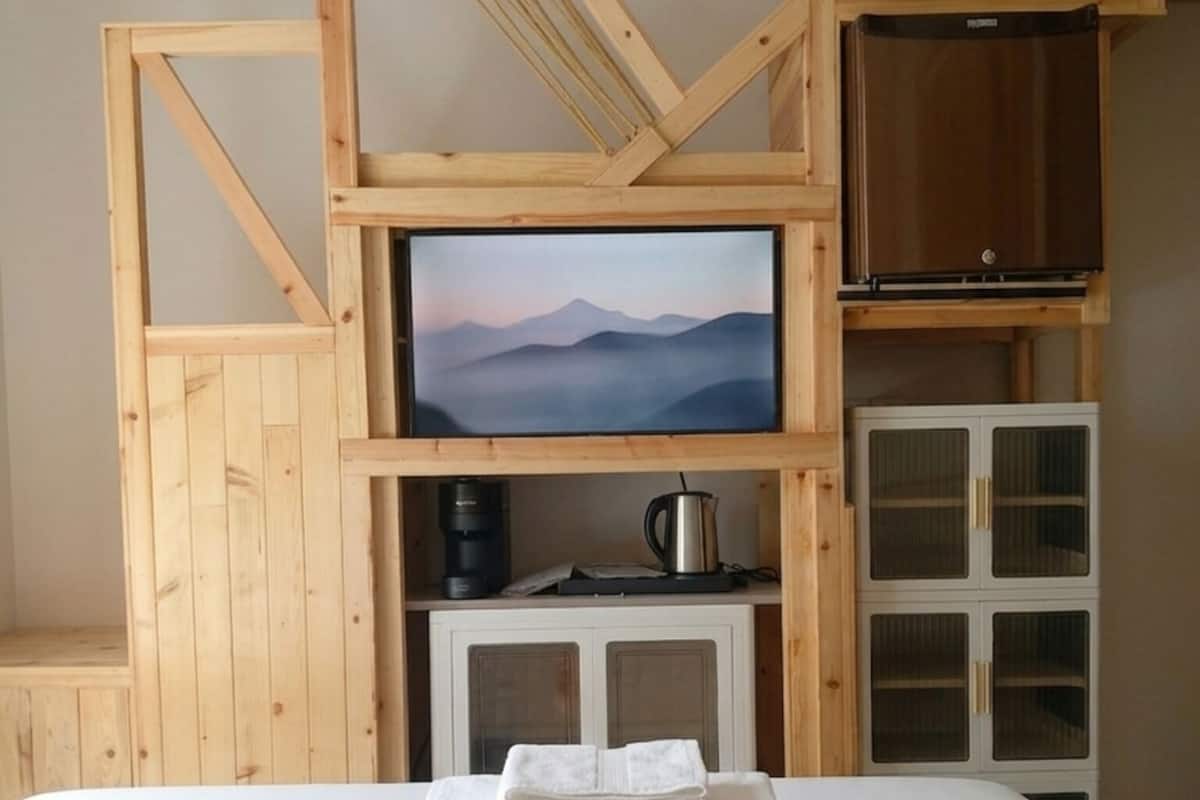 A room with a bed, a television mounted on a wooden wall, a coffee maker, and a cabinet with glass doors.